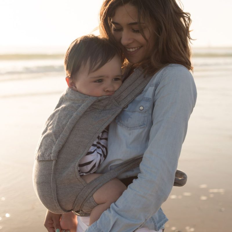 Maman portant son bébé en porte-bébé physiologique sur la plage
