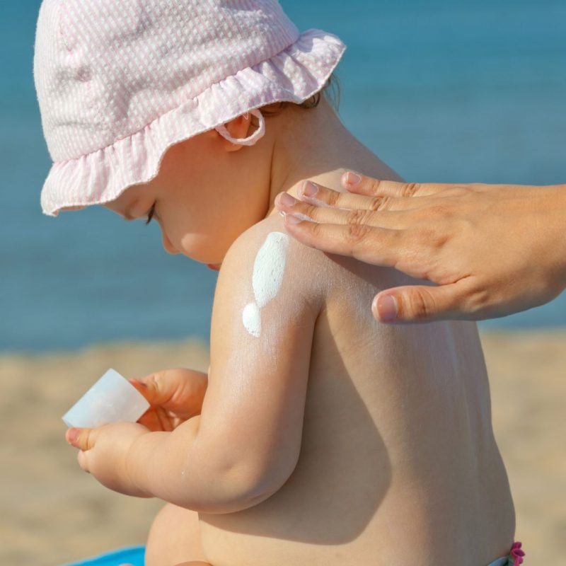 Application de crème solaire sur l’épaule d’un bébé avec un chapeau à la plage