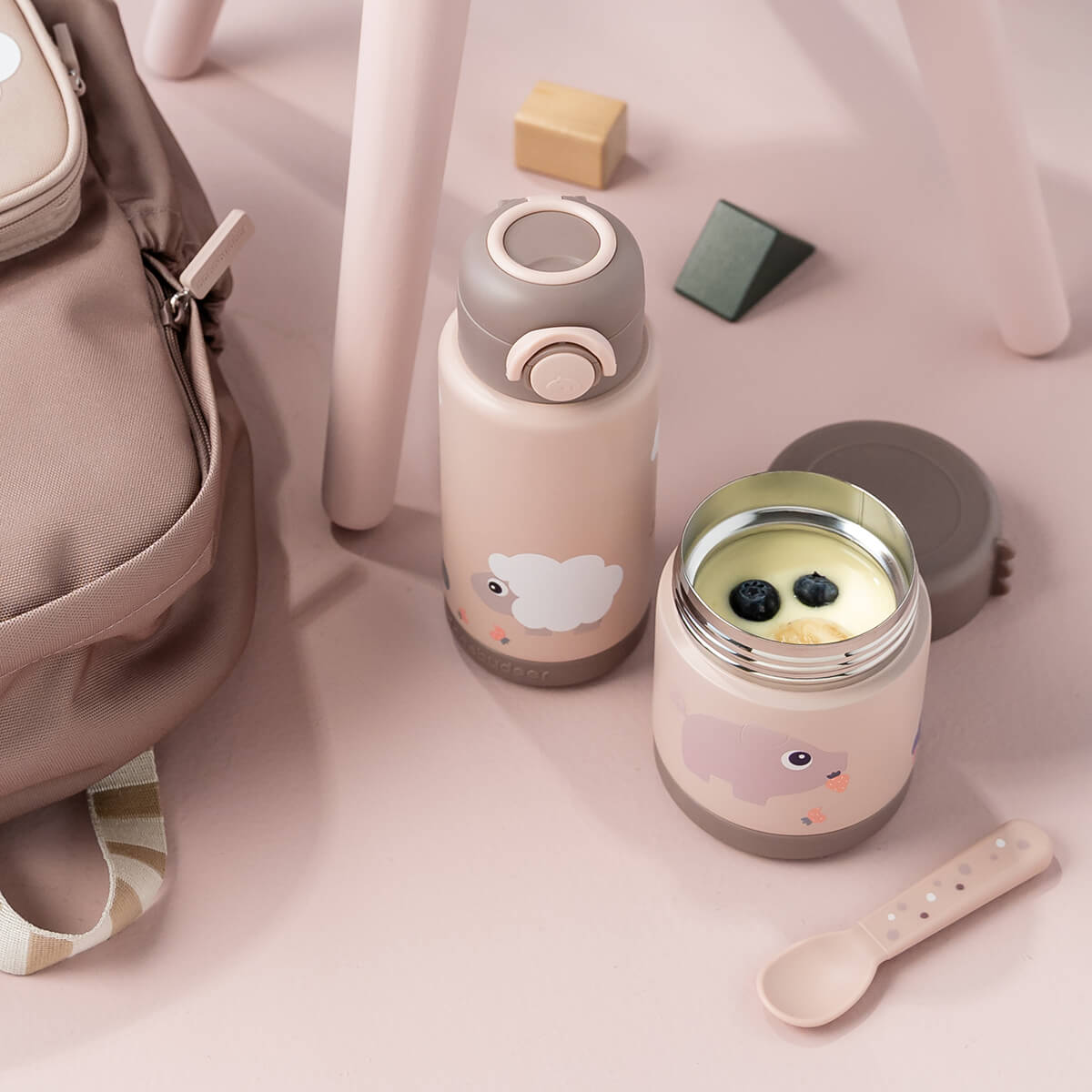Gourde isotherme Done By Deer rose et pot assorti posés à côté d’un sac et d’une cuillère sur un sol rose