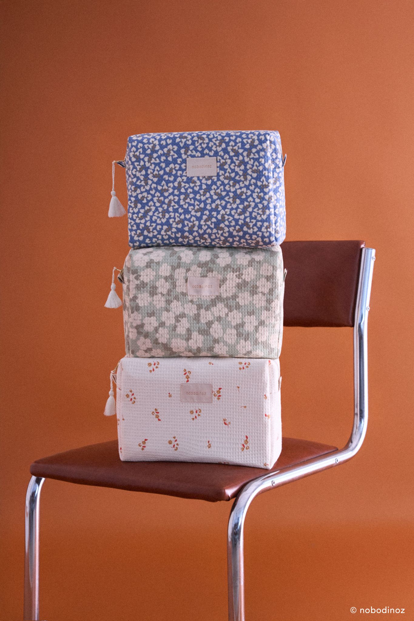 Trousse de toilette Nobodinoz Diva en version Blue Yumiko, Green Tea et motifs floraux, empilées sur une chaise vintage