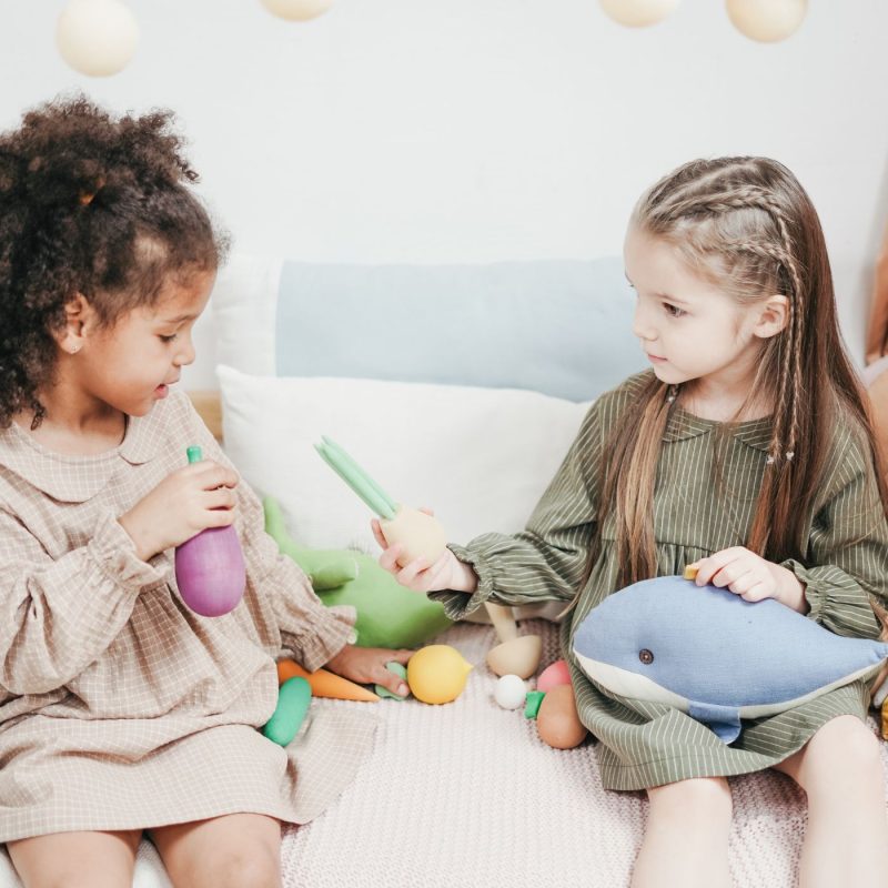 Deux petites filles échangent des légumes en bois en jouant sur un lit, dans une scène de partage et de jeu symbolique.