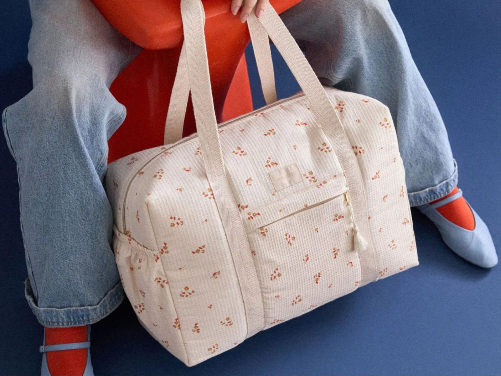 Sac à langer Nobodinoz Yellow Lily à motifs fleuris posé entre les jambes d’une personne assise, sur fond bleu