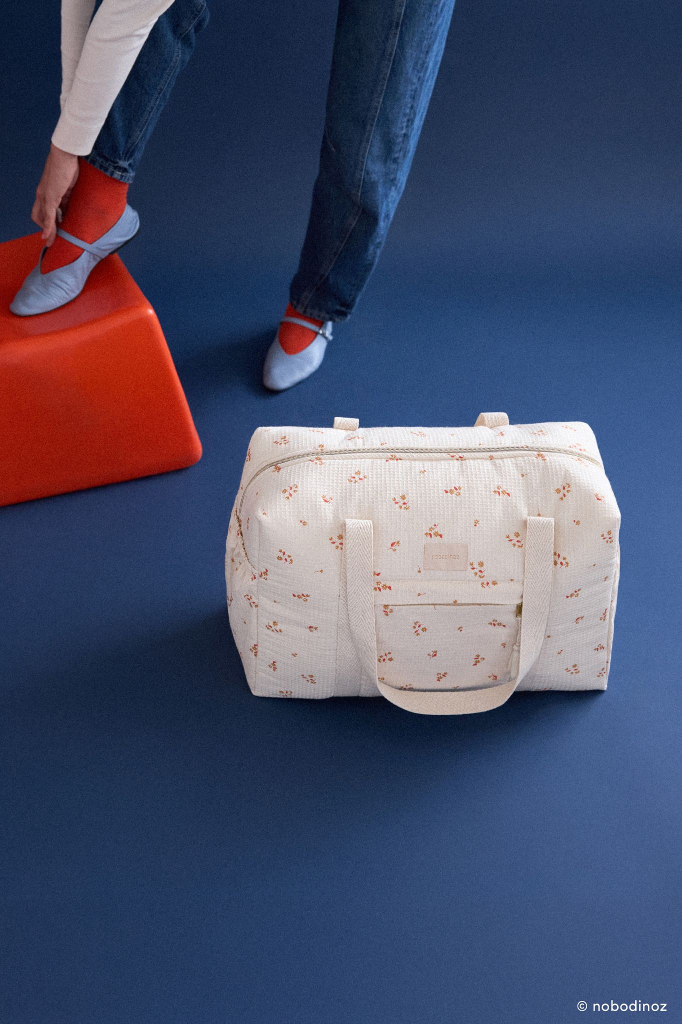 Sac à langer imperméable Opera yellow lily Nobodinoz posé au sol à côté d’une personne