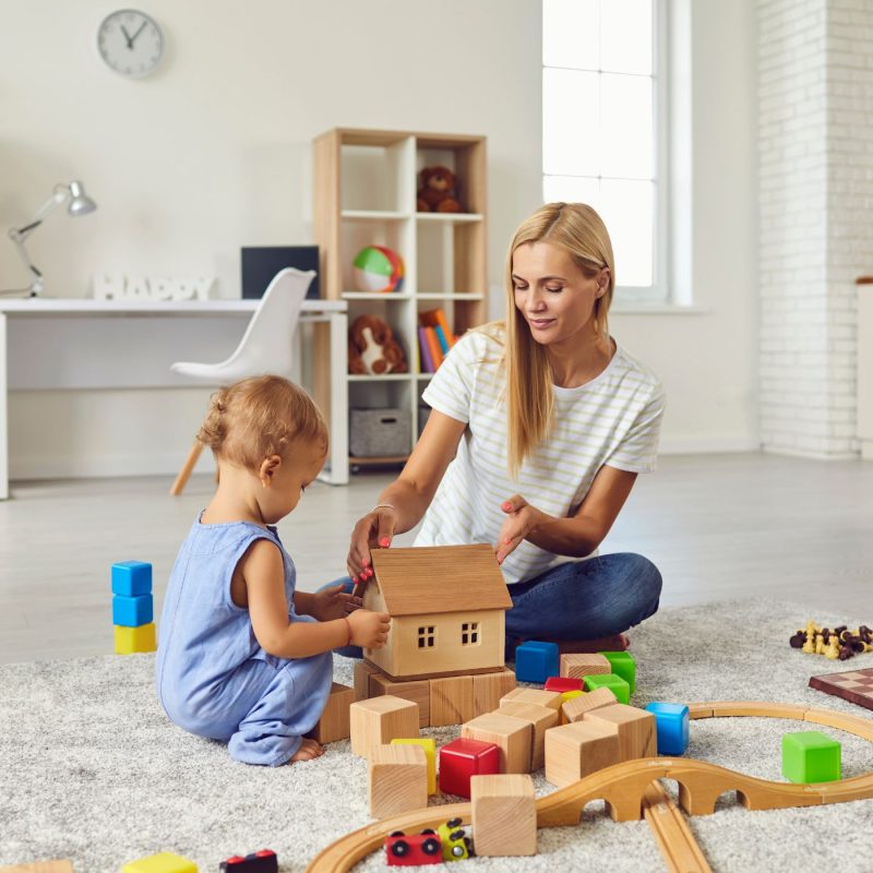 Babysitter jouant avec un enfant au sol avec des jouets en bois, exemple de garde réussie.