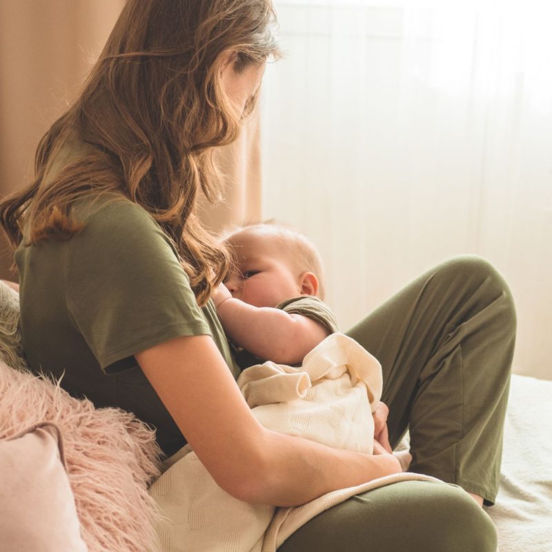 Jeune maman allaitant son bébé, assise sur un lit, en post-partum.
