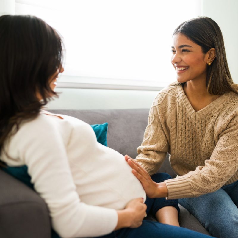 Une sage-femme souriante accompagne une femme enceinte assise sur un canapé, illustrant l’importance du suivi postnatal et du soutien émotionnel.