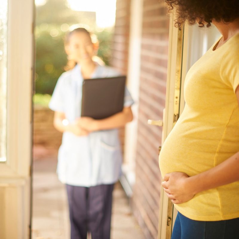 Une sage-femme arrive au domicile d’une jeune maman pour une visite postnatale, illustrant le suivi médical à domicile après l’accouchement.