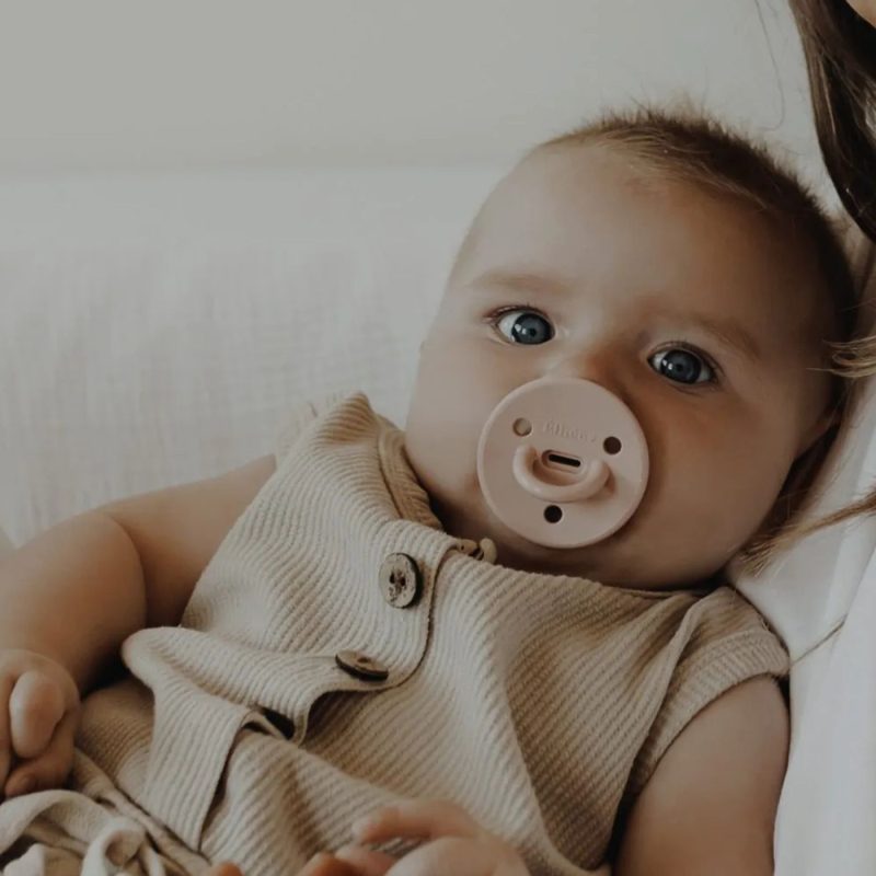 Bébé allongé dans les bras de sa maman, portant une sucette Élhée rose poudré